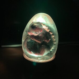 Könnte beinhalten: Eine klare, eiförmige Lampe aus Glas mit einer roten und schwarzen Eidechsenfigur im Inneren. Die Lampe ist eingeschaltet und leuchtet grün.