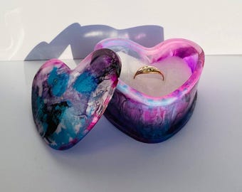 Può includere: Una scatola per anelli a forma di cuore con un coperchio. La scatola è realizzata in resina e presenta un motivo a spirale di rosa, blu, nero e bianco. Il coperchio è aperto e all'interno c'è un anello d'oro.