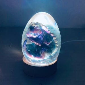 Könnte beinhalten: Ein klares, eiförmiges Nachtlicht aus Glas mit einer kleinen, schillernden Drachenfigur im Inneren. Das Licht beleuchtet den Drachen und erzeugt einen magischen Schein.