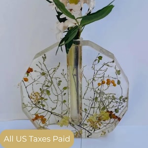 Könnte beinhalten: Transparente Harzvase mit achteckiger Form, mit eingebetteten getrockneten Blumen und Blättern. Die Vase enthält einen Stiel mit weißen Blüten und grünen Blättern. Der Text "All US Taxes Paid" befindet sich unten.