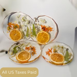 Puede incluir: Cuatro posavasos redondos de resina transparente con flores secas y rodajas de naranja incrustadas. Los posavasos tienen un borde facetado. Las flores son naranjas, blancas y verdes. Las rodajas de naranja son de color naranja seco.