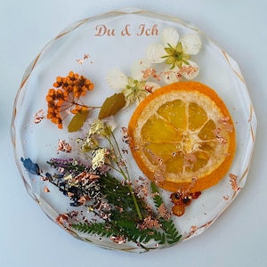 Könnte beinhalten: Ein klarer, runder Harz-Untersetzer mit einer getrockneten Orangenscheibe, Blumen und kupferfarbenen Flocken. Der Untersetzer trägt den Text "Du & Ich" in Kupferfarbe. Der Untersetzer ist ein dekorativer Artikel.