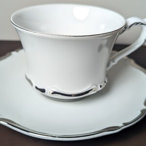 May include: A white porcelain teacup and saucer set with a silver trim. The cup has a rounded shape and a handle. The saucer has a scalloped edge.