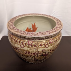 Puede incluir: Un cuenco de cerámica decorativo con un patrón floral dorado y rojo. El cuenco tiene un borde ancho y una base redondeada. Presenta un diseño pintado de dos peces dorados nadando en el agua.
