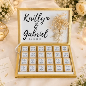 K&ouml;nnte beinhalten: Eine gold umrandete rechteckige Schachtel mit vierundzwanzig quadratischen Pralinen, jeweils in Goldfolie gewickelt mit einem wei&szlig;en Etikett, bedruckt mit den Namen Kaitlyn & Gabriel und dem Datum 05.21.2026. Der Deckel zeigt dieselben Namen und getrocknete Blumen.