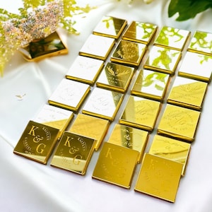 May include: A collection of square gold-toned acrylic favors arranged in a grid. Each piece features custom laser-engraved text including the initials K&G, the names Kaitlyn and Gabriel, and the date 04.04.25, alongside various floral motifs.
