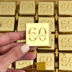 May include: Gold-coloured square chocolates arranged in a tray. Each chocolate is engraved with the number "60" and the words "Kami's Birthday 12.07.24". One chocolate is held up, showcasing the details. The chocolates are a celebratory treat.