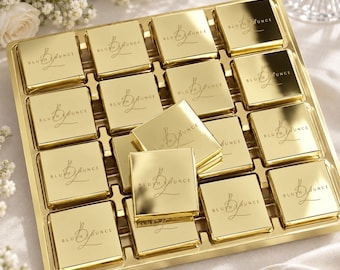 Op de afbeelding: Een goudkleurig dienblad met zestien vierkante, metallic gouden items, elk voorzien van de tekst 'BLUSH LOUNGE' en een gestileerd 'BL'-monogram. De items zijn in een raster geplaatst, waarbij één item schuin bovenop ligt.