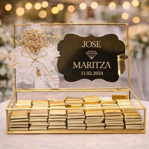 May include: A gold-framed glass box filled with gold-wrapped chocolates. The box features a decorative label with the names "JOSE" and "MARITZA" and the date "11.02.2024". A small bouquet and bow adorn the top.