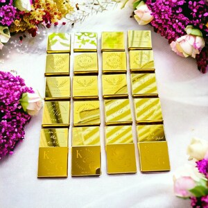 Op de afbeelding: Goudkleurig verpakte chocolade gunsten gerangschikt op een wit oppervlak, met bloemstukken op de achtergrond. Elke chocolade vierkant is gepersonaliseerd met initialen, namen en data, ideaal voor bruiloften of speciale evenementen.