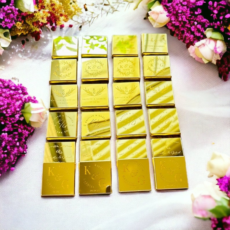 Op de afbeelding: Goudkleurig verpakte chocolade gunsten gerangschikt op een wit oppervlak, met bloemstukken op de achtergrond. Elke chocolade vierkant is gepersonaliseerd met initialen, namen en data, ideaal voor bruiloften of speciale evenementen.