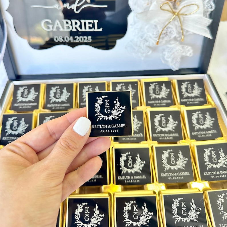 以下が含まれることがあります： 結婚式用のパーソナライズされたラベルが付いたチョコレートの箱。チョコレートは正方形で、金色のホイルで包まれ、カップルのイニシャル、名前、結婚式の日付が記載された黒いラベルが付いています。Kaitlyn & Gabriel, 08.04.2025.