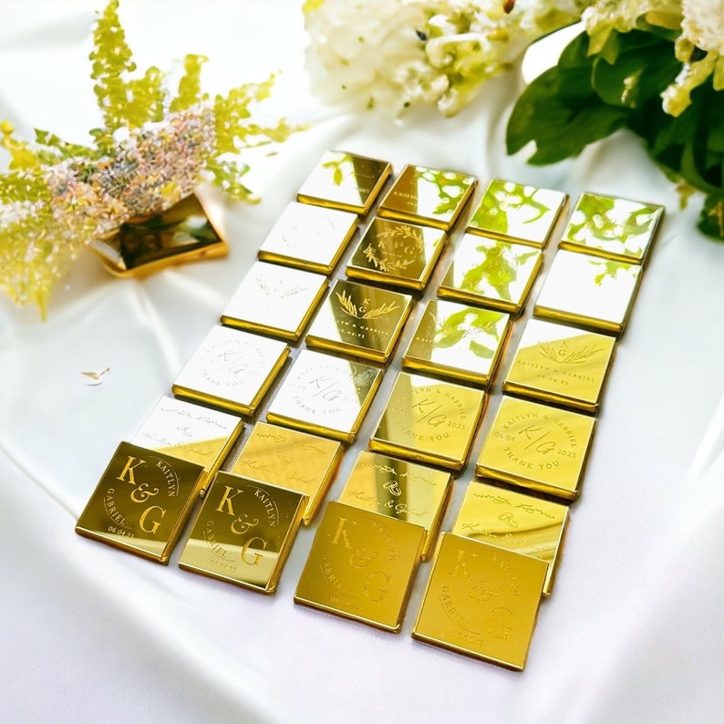 May include: Arrangement of gold-colored, square-shaped favors with personalized text and initials. The favors are arranged on a white satin surface, with floral accents in the background. The text includes names, dates, and the words "Thank You".