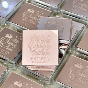 May include: Square, rose-gold favors with the text "Mis Quince A&ntilde;os" and the name "Violeta" along with the date "27 diciembre 2024" engraved on the surface. The favors are in clear plastic containers.