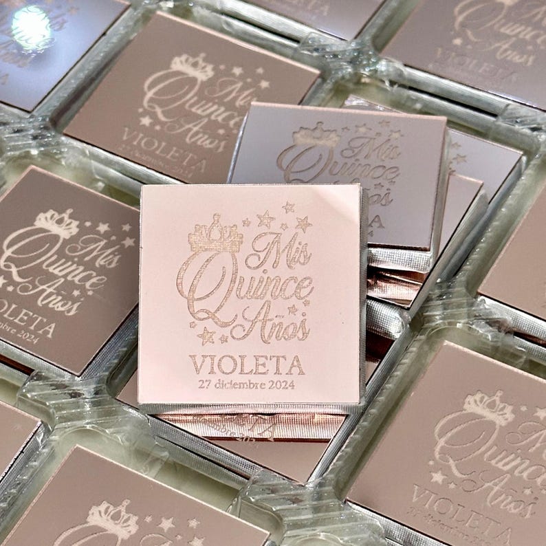 May include: Square, rose-gold favors with the text "Mis Quince A&ntilde;os" and the name "Violeta" along with the date "27 diciembre 2024" engraved on the surface. The favors are in clear plastic containers.