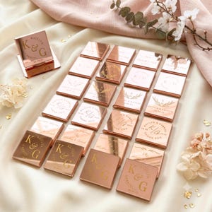 May include: A collection of square rose gold mirror acrylic favors, each engraved with custom text including names like Kaitlyn and Gabriel, initials K and G, and dates. One piece stands upright on a small base, while others lie flat in a grid arrangement.