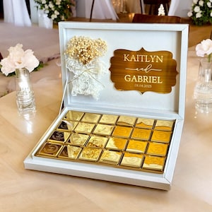 以下が含まれることがあります： 金色のチョコレートが詰められた白い箱。金色のプレートには「KAITLYN and GABRIEL」と日付「08.04.2025」が刻まれています。箱はドライフラワーとレースで飾られています。