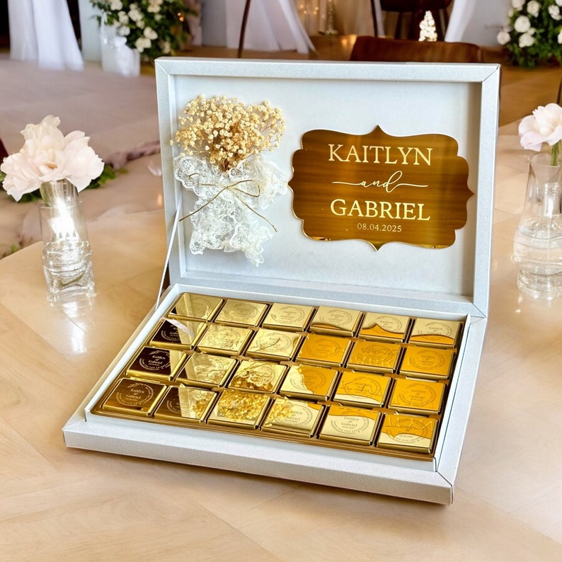 以下が含まれることがあります： 金色のチョコレートが詰められた白い箱。金色のプレートには「KAITLYN and GABRIEL」と日付「08.04.2025」が刻まれています。箱はドライフラワーとレースで飾られています。