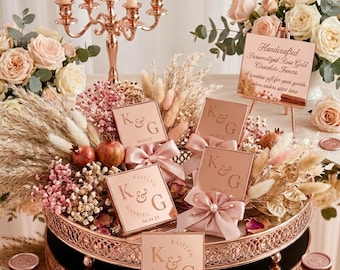 May include: A round rose gold metal tray holding square chocolate favors with the initials K&G and the date 06.04.23. The favors are decorated with pink satin bows, dried pampas grass, and small pomegranates, displayed near a rose gold candelabra.
