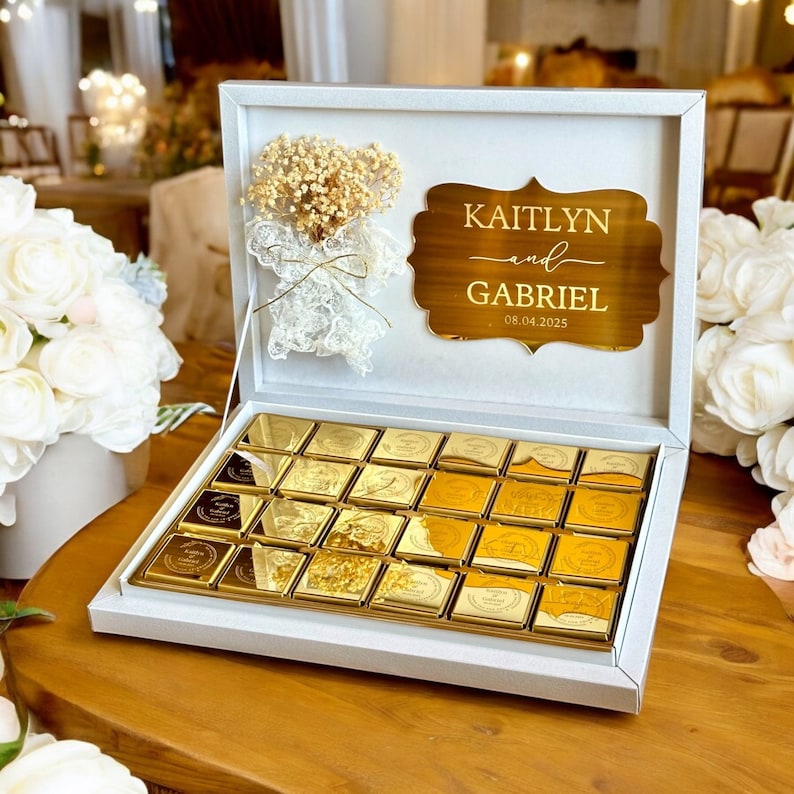 以下が含まれることがあります： 金色のチョコレートが詰められた白い箱。箱には「Kaitlyn and Gabriel」の名前と日付「08.04.2025」が刻まれた金色のプレートが付いています。箱はドライフラワーのアレンジメントとレースで飾られています。チョコレートは箱の中にきれいに並べられています。