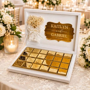 May include: A white rectangular gift box containing twenty-four square gold-toned chocolate wrappers. The inside lid features a gold plaque with the text KAITLYN and GABRIEL 08.04.2025 and a decorative arrangement of dried white flowers and lace.