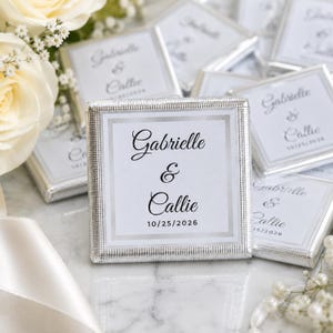 K&ouml;nnte beinhalten: Eine quadratische Schokoladentafel in silberner Folie mit einem wei&szlig;en Etikett, das den schwarzen Aufdruck Gabrielle & Callie und das Datum 10/25/2026 tr&auml;gt. Das Etikett hat einen silbernen Rand und die Folie ein strukturiertes Gittermuster.