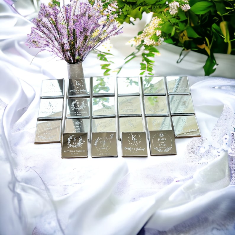 May include: A collection of silver square favors, each engraved with names, initials, and dates. The favors are arranged on a white surface, with a bouquet of purple flowers and greenery in the background.