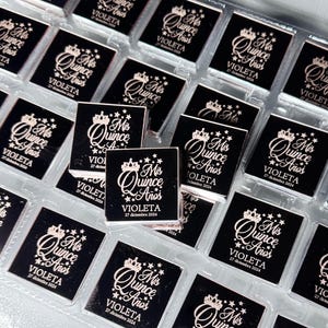 May include: A collection of small, square, black favors with a decorative design. Each favor features the text "Mis Quince A&ntilde;os" in a stylized font, along with the name "Violeta" and the date "27 diciembre 2024". The design includes a crown and stars.