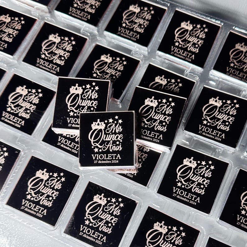 May include: A collection of small, square, black favors with a decorative design. Each favor features the text "Mis Quince A&ntilde;os" in a stylized font, along with the name "Violeta" and the date "27 diciembre 2024". The design includes a crown and stars.