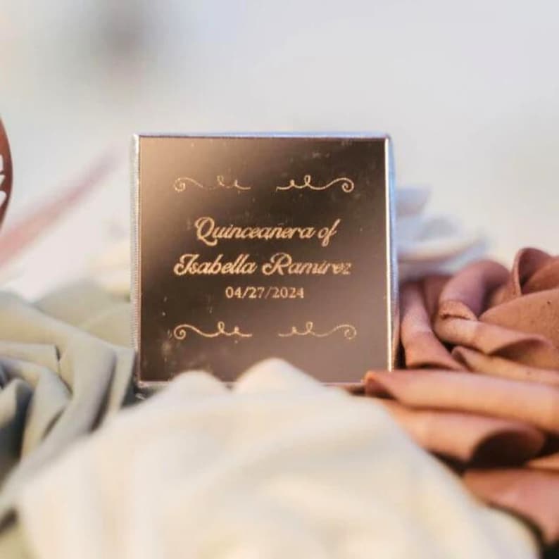 May include: A square, rose gold box with engraved text reading "Quinceanera of Isabella Ramirez 04/27/2024". The box is surrounded by soft, folded fabric in shades of white, gray, and rose.