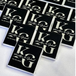 May include: A collection of black square favors with silver lettering. Each favor has the initials "KG", the names "Kaitlyn & Gabriel", and the date "04.08.2025". The favors are arranged on a white surface.