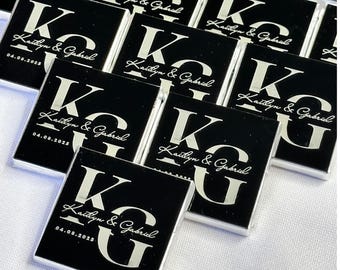 May include: A collection of black square favors with silver lettering. Each favor has the initials "KG", the names "Kaitlyn & Gabriel", and the date "04.08.2025". The favors are arranged on a white surface.