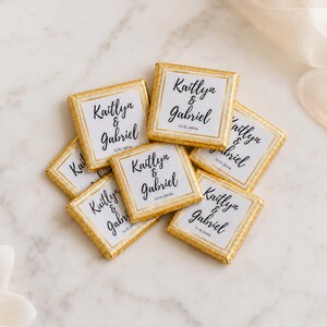 K&ouml;nnte beinhalten: Eine Ansammlung quadratischer Schokoladentafeln in Goldfolie. Jede Tafel hat ein wei&szlig;es Etikett mit goldenem Rand, bedruckt mit den Namen Kaitlyn & Gabriel in schwarzer Schreibschrift und dem Datum 11/21/2026 in einer einfachen serifenlosen Schrift.