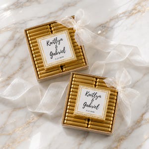 K&ouml;nnte beinhalten: Zwei quadratische Geschenkboxen gef&uuml;llt mit kleinen Schokoriegeln in gl&auml;nzender Goldfolie. Jede Box hat ein wei&szlig;es Etikett mit Goldrand, bedruckt mit den Namen Kaitlyn & Gabriel und dem Datum 11.21.2026. Ein transparentes wei&szlig;es Band ist zu einer Schleife gebunden.