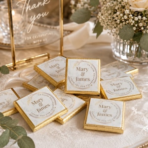K&ouml;nnte beinhalten: Quadratische Schokoladenpr&auml;sente in wei&szlig;em Papier mit goldener Folienkante. Jedes St&uuml;ck zeigt ein kreisf&ouml;rmiges Design mit goldenen Lorbeerzweigen und dem Text Mary & James Thank You For Celebrating With Us auf einem wei&szlig;en Marmormuster.