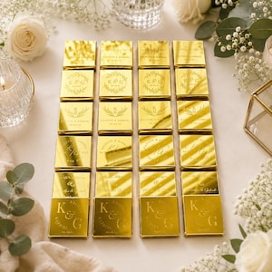 May include: A collection of twenty square, gold-toned metallic favours arranged in a grid. Each piece features engraved text and botanical illustrations, including the initials K & G, the names Kaitlyn & Gabriel, and the date 09.09.23.