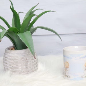May include: White ceramic mug with a blue and white design featuring two angels. The mug is sitting on a white fluffy surface next to a potted green succulent plant.