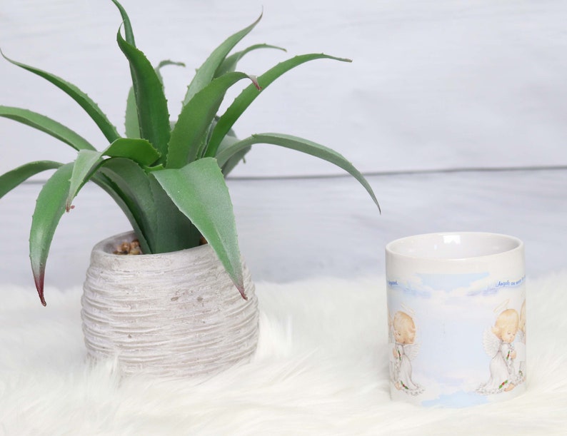 May include: White ceramic mug with a blue and white design featuring two angels. The mug is sitting on a white fluffy surface next to a potted green succulent plant.