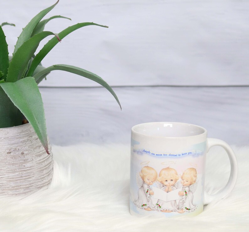 May include: White ceramic mug with a blue and white illustration of three angels singing. The text on the mug reads "Angels are never too distant to hear you."