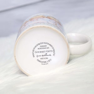 May include: White ceramic mug with a blue and white design featuring a couple of angels. The mug has a black and white label on the bottom that reads "Gourmet Gifts from Hallmark" and "Dishwasher and Microwave Safe".