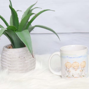 May include: White ceramic mug with a blue and white design featuring three angels singing. The text on the mug reads "All angels come to us disguised".