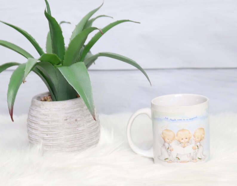 May include: White ceramic mug with a blue and white design featuring three angels singing. The text on the mug reads "All angels come to us disguised".