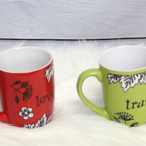 May include: Four ceramic mugs with floral designs and inspirational words. The mugs are blue, red, green, and yellow. The words on the mugs are "faith", "love", "trust", and "joy".