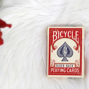 May include: A red and white playing card deck box with the word "BICYCLE" in a white arch over a blue spade playing card. The text "RIDER BACK PLAYING CARDS" is printed below the spade.