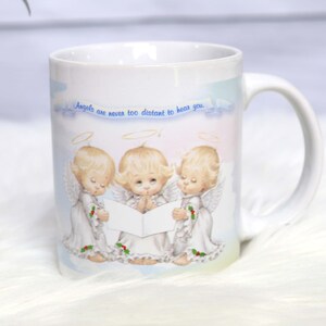 May include: White ceramic mug with a colorful design of three angels singing. The mug has the text "Angels are never too distant to hear you."