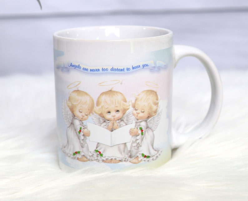 May include: White ceramic mug with a colorful design of three angels singing. The mug has the text "Angels are never too distant to hear you."