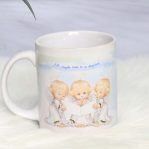 May include: White ceramic mug with a blue and white illustration of three angels singing. The mug has the text "All Angels come to us disguised" printed on the front.