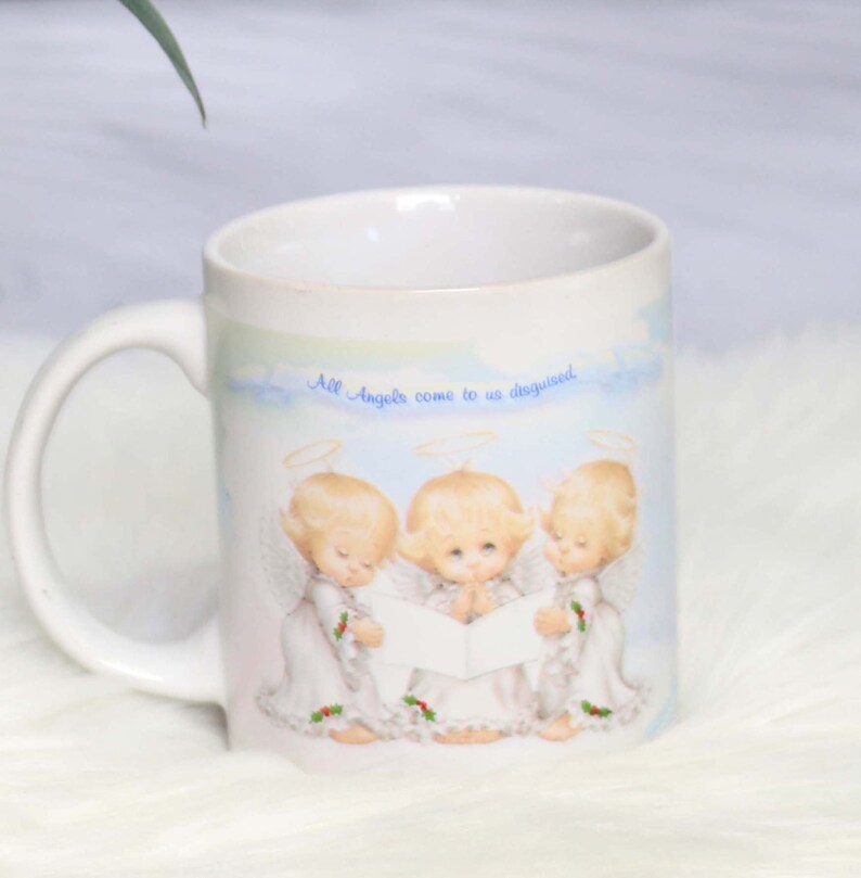 May include: White ceramic mug with a blue and white illustration of three angels singing. The mug has the text "All Angels come to us disguised" printed on the front.