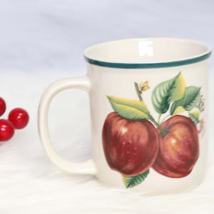 May include: Cream-colored ceramic mug with a green rim and handle. The mug features a detailed illustration of two red apples with green leaves and small white flowers. A decorative item for the kitchen.