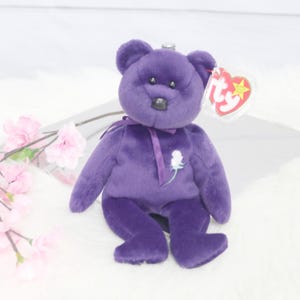 May include: A plush purple teddy bear with a white rose embroidered on its chest. The bear has a black nose, small black eyes, and a red heart-shaped tag. The bear is sitting on a white surface.